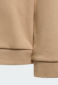 Béžová mikinová tkanina zobrazujúca textúrovaný povrch, rebrované manžety a detaily švov. Materiál pôsobí mäkko a odolne.