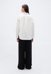 Λευκό πουκάμισο με μακριά μανίκια και κουμπιά μπροστά, με λεπτομέρεια yoke στην πλάτη, σε συνδυασμό με μαύρο παντελόνι φαρδιάς γραμμής. Λείο ύφασμα με κλασικό γιακά.