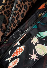 A steppelt kabát belsejében barna leopárdminta és fekete anyag található színes hal- és virágmintákkal, melyeken cipzárok és tépőzárak láthatók.