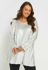 Skinande silverfärgad långärmad topp med rund halsringning, texturerad tyg och utsvängd design, kombinerad med svarta leggings.