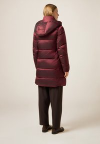 Bordeauxfarvet vatteret jakke med hætte, quiltet design og en skinnende tekstur. Båret over løstsiddende brune bukser og sort fodtøj.