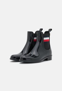 Bottines en caoutchouc noires avec une finition brillante, panneaux latéraux élastiques dotés de rayures rouge, blanc et bleu, et un petit talon.