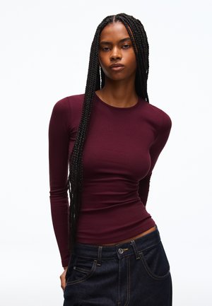 Ung kvinde med langt fletning iført en tætsiddende mørkerød bluse med lange ærmer og mørkeblå jeans mod en hvid baggrund.