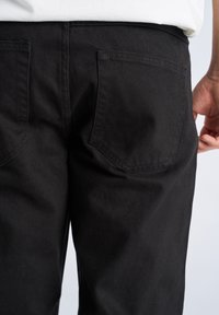 Pantaloni in denim nero con vestibilità classica, dotati di due tasche posteriori. Il tessuto ha una texture liscia e cuciture robuste lungo le cuciture.