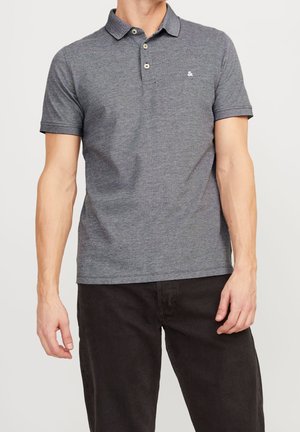 Man wearing a short-sleeve gray polo shirt with two buttons and small logo on chest, paired with black pants against plain background.