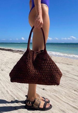 Persona con costume da bagno blu che tiene una grande borsa marrone intrecciata su una spiaggia sabbiosa con oceano e cielo sereno sullo sfondo.