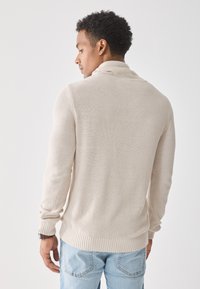 Pull en tricot beige avec un col haut et des poignets côtelés, associé à un jean bleu clair. La texture est douce et lâchement tissée.
