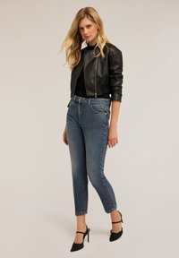Czarna skórzana kurtka bombowiec, dopasowana czarna bluzka, jasnoniebieskie dżinsy prostego kroju i czarne buty na obcasie. Modelka prezentuje zrelaksowaną pozę.