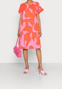 Vestido naranja brillante con patrones geométricos rosas, mangas con volantes y falda de varios niveles. Combinado con mocasines de tacón rosa y un bolso a juego.