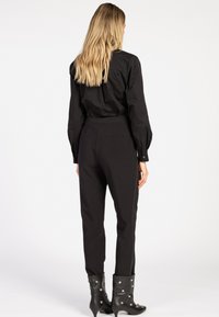 Vrouw met lang blond haar die een zwart langoor-t-shirt, zwarte strakke broek met zijstuds en zwarte enkellaarzen met zilveren studs draagt.