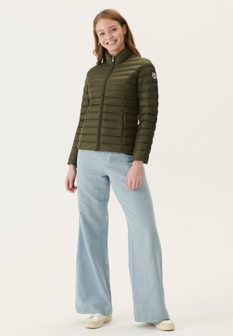 Groene pufferjack met een hoge kraag, ritsluiting en smalle horizontale naden. Gepaard met lichtblauwe broek met brede pijpen en witte sneakers.