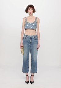 Denim crop-top med sølvdetaljer, kombinert med høytlivede blå jeans. Modellen holder en gullklynge og har på seg svarte hælede sandaler.