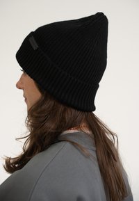 Berretto lavorato a maglia nero con una trama a coste, caratterizzato da un dettaglio etichetta discreto, che si affina a una punta nella parte superiore. Indossato da una persona con capelli lunghi.