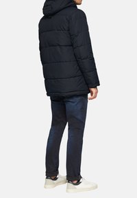 Marineblauwe pufferjack met horizontale stiksels, afneembare capuchon, gedragen over donkere spijkerbroeken en lichte sneakers, ontworpen voor warmte en comfort.