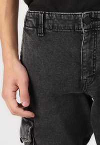 Černé džínové cargo kalhoty s přední kapsou a kontrastním bílým šitím. Tkanina vypadá texturovaně s mírným leskem.