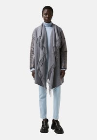 Šedý šátek s třásněmi, který je vzatý přes světlomodrý rolák a vycpanou bundu, doplněný světlomodrými kalhotami a černými robustními botami.