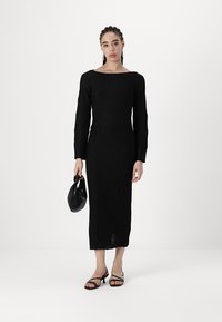 Vestido preto de manga longa com padrão geométrico texturizado, silhueta ajustada e comprimento até a metade da panturrilha. Combinado com uma pequena bolsa preta e sandálias minimalistas.