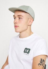 Gorra de pana verde claro con visera plana, combinada con una camiseta blanca que presenta un diseño impreso en verde en el lado izquierdo del pecho. Mangas cortas.
