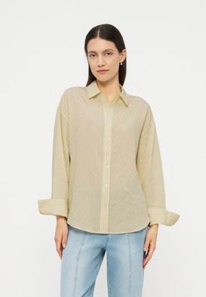 Kobieta o długich ciemnych włosach, ubrana w jasnobeżową, przezroczystą, paskowaną koszulę na guziki oraz jasnoniebieskie dżinsy, stojąca na tle jednolitego białego tła.