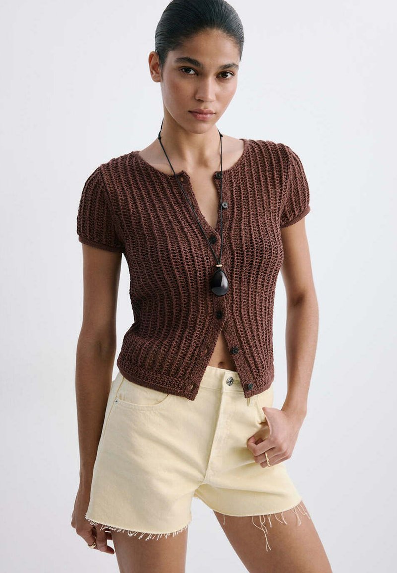 Femme portant un haut marron en maille à manches courtes boutonné et un short crème à ourlet effiloché, avec un collier pendentif noir, posant devant un fond uni.