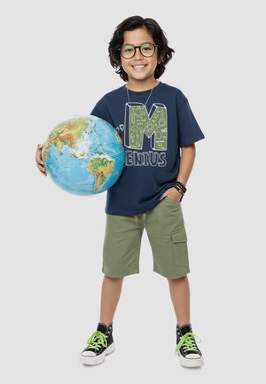 Usměvavé dítě s brýlemi, v námořnicky modrém tričku s nápisem „Genius“ a zelených kraťasech drží globus ukazující kontinenty na světlém pozadí.