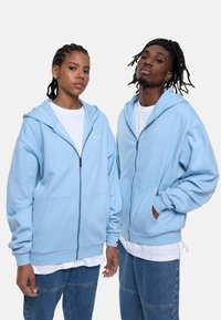 Light blue zip-up hoodies made of cotton blend fabric, featuring a front pocket and ribbed cuffs. Worn over white t-shirts and paired with denim.