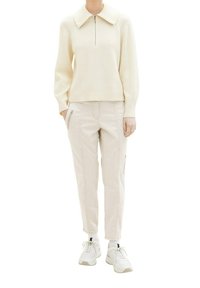 Personne portant un haut en maille crème à manches longues avec col, un pantalon cargo beige avec poches zippées, des chaussettes blanches et des baskets blanches.