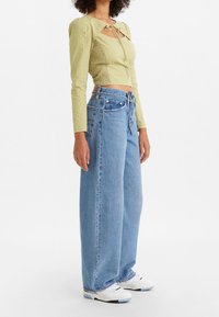 Ljusgrön långärmad topp med strukturerat mönster och nyckelhål fram; kombinerad med lösa, ljusblå denimjeans och vita sneakers.