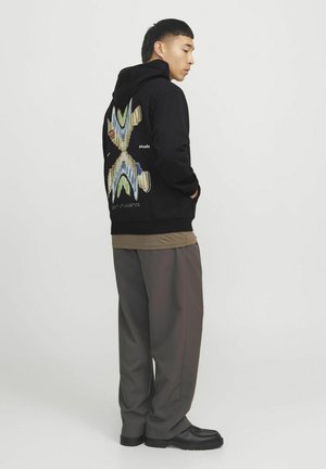 Schwarzer Kapuzenpullover mit einem bunten Grafikdesign auf dem Rücken, kombiniert mit lockeren grauen Hosen und schwarzem Schuhwerk, was Komfort und lässigen Stil betont.