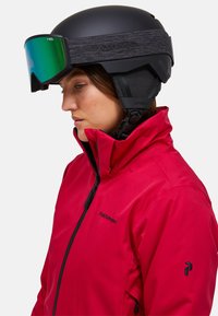 Vrouw met een zwarte skhelmet, groene goggles en een rode Peak Performance jas, kijkt naar beneden naar links.