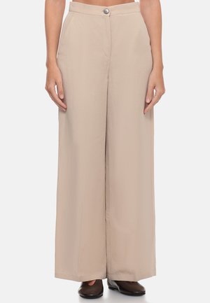 Pantaloni beige a vita alta e gamba larga con chiusura con bottone e cerniera, indossati con scarpe marrone scuro con tacco basso.