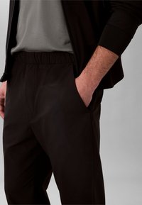 Mees, kes kannab musti pükse elastse vöökohaga ja hoiab käed taskutes, kombineeritud halli särgi ja musta jopetega.