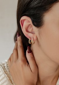 Pendientes de aro dorados con un diseño retorcido, colgando de la lóbulos, acompañados de una mano tocando suavemente la oreja. Piel suave y cabello oscuro visibles.