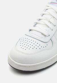 White leather sneaker with perforated toe box, flat white laces, padded tongue, and a textured rubber sole with black accent.