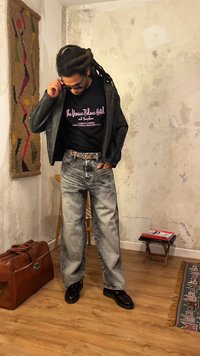 Άνδρας με ντραντλοκς ρυθμίζει γυαλιά ηλίου, φοράει μαύρο δερμάτινο μπουφάν, μαύρο T-shirt με τύπωμα, τζιν με οξύ πλύση και μαύρα παπούτσια σε ένα ρουστίκ δωμάτιο.