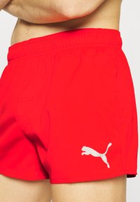 Rote Schwimmshorts mit elastischem Bund, ausgestattet mit einer kleinen Seitentasche und einem weißen Puma-Logo auf der unteren linken Seite. Glatte Textur.