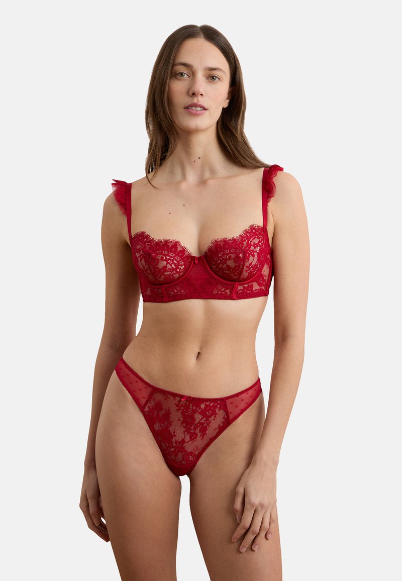 Femme portant un soutien-gorge en dentelle rouge avec bretelles volantées et une culotte haute en dentelle transparente assortie, debout devant un fond blanc uni.