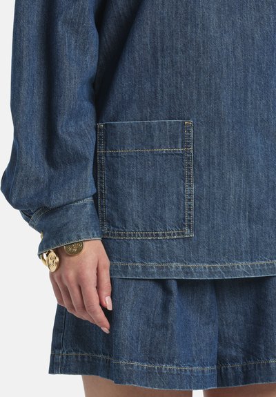Main reposant sur le côté, portant un haut en denim à manches longues avec une grande poche carrée et une jupe en denim assortie, ornée d'un bracelet à breloques doré.