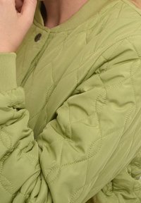 Nærbillede af en person iført en lys grøn quiltet jakke med ribbet krave og manchetter, som holder hånden tæt på ansigtet.