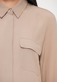 Bluza bej dintr-un material ușor, cu guler clasic, închidere frontală cu nasturi și buzunar pe partea stângă. Textură netedă.