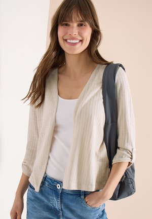 Mladá žena se usmívá, nosí béžový otevřený kardigan, bílý top, modré džíny a má přes jedno rameno tmavě šedý batoh.