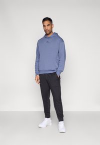 Μπλε φούτερ με κουκούλα και λογότυπο μπροστά, συνδυασμένο με μαύρα αθλητικά παντελόνια και λευκά αθλητικά παπούτσια. Λείο ύφασμα, άνετη εφαρμογή και ριμπ μανσέτες.