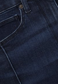 Sötétkék farmeranyag látható varrásokkal, elülső zsebbel és fém szegecs részlettel a farmerekon.