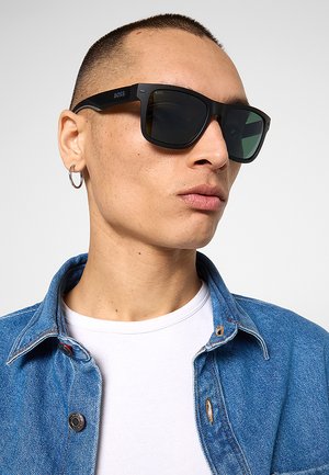 Uomo con capelli corti che indossa occhiali da sole rettangolari neri, un orecchino a cerchio, una camicia bianca e una giacca di jeans blu.