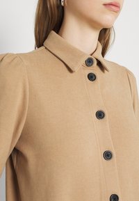 Camicia beige con bottoni, dotata di colletto, bottoni neri e maniche a sbuffo, realizzata in un tessuto testurizzato.