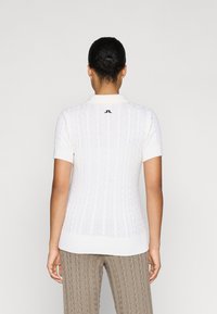 Polo corto a maniche corte di colore bianco, con motivo a treccia e orlo a coste. Dettaglio con logo nero sul colletto posteriore.