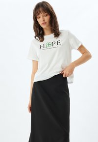 Hvid bomulds t-shirt med "HOPE" og et grønt kløvergrafik, parret med en sort, lang, svævende nederdel med glat tekstur.