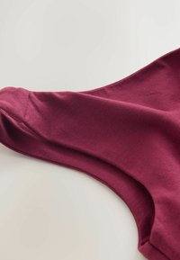 Burgundijas kokvilnas bezpieģe ar mīkstu tekstūru un noapaļotu kakla izgriezumu, ar gludu šuvi gar roku atveri un ķermeni.