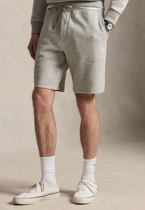 Homme portant un short gris clair à cordon, des chaussettes blanches montantes, des baskets blanches et un sweat-shirt gris avec une montre au poignet gauche.