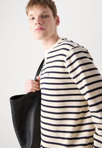 Stripet bomullsskjorte med lang erme i krem med marineblå striper, med rund halsutringning og brodert logo. En svart lær totebag holdes i hånden.